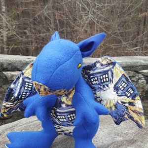 May include: Blue plush dragon with wings made of fabric featuring a Starry Night-inspired print. The dragon has a yellow eye and is sitting on a grey stone surface.