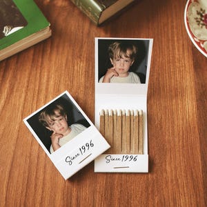 May include: Two white matchbooks featuring a photograph of a young child on the cover and inside flap. Each matchbook is printed with the text Since 1996 in a black script font. One matchbook is open, revealing a row of light brown matches.