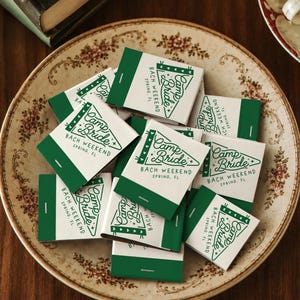 May include: A collection of rectangular matchboxes with a white and dark green color scheme. Each box features a graphic of a pennant flag with the text Camp Bride BACH WEEKEND SPRING, FL printed in dark green ink on the white surface.