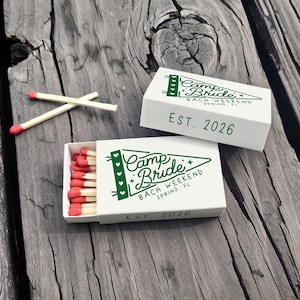 May include: White matchboxes with green text that reads "Camp Bride Bach Weekend Spring, FL EST. 2026". One box is open, revealing red-tipped matches. Two matches are placed on the wooden surface.