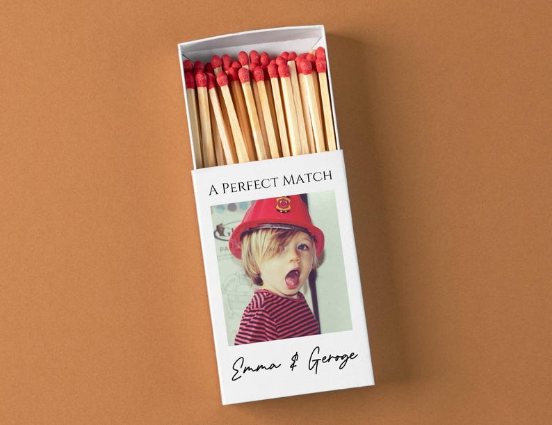 May include: A white matchbox with the text "A PERFECT MATCH" and a photo of a child wearing a red firefighter hat. The box is filled with wooden matches with red tips. The names "Emma & George" are written below the photo.