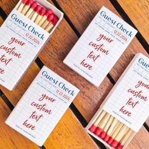 May include: Four white matchboxes with a grid pattern and the text "Guest Check" and "your custom text here" in red. Two boxes are open, revealing red-tipped matches. The date "12.22.2026" is printed on the boxes.