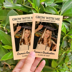 May include: Two rectangular tan paper seed packets labeled GROW WITH ME WILDFLOWER SEEDS. Each packet features a photograph of a graduate in a cap and gown, with the text Emma's Graduation CLASS OF 2027 printed in black at the bottom of the packet.
