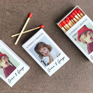 May include: Three white matchboxes with custom photos and text. Two matches are crossed on the surface. The matchboxes have a photo of a child wearing a red hat and striped shirt. The text on the boxes reads "A PERFECT MATCH" and "Emma & Geroge".