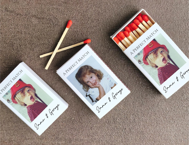 May include: Three white matchboxes with custom photos and text. Two matches are crossed on the surface. The matchboxes have a photo of a child wearing a red hat and striped shirt. The text on the boxes reads "A PERFECT MATCH" and "Emma & Geroge".