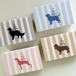 May include: Four rectangular boxes with striped patterns in gray, blue, tan, and pink. Each box features a dog silhouette carrying a stack of champagne glasses. The boxes are ideal for small gifts or party favors.