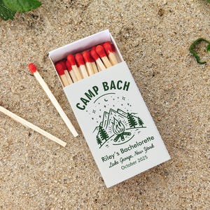 May include: A white matchbox with the text "CAMP BACH" and a green mountain and campfire design. The match heads are red. Two matches are lying on the sand next to the box. The text "Riley's Bachelorette Lake George, New York October 2025" is also on the box.