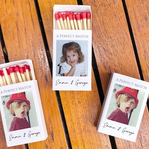 May include: Three white matchboxes with red-tipped matches. Each box features a photo of a child and the text "A PERFECT MATCH" and the names "Emma & George". The boxes are on a wooden surface.