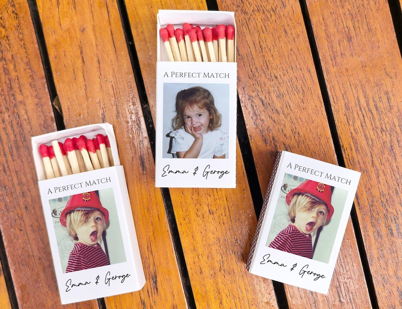 May include: Three white matchboxes with red-tipped matches. Each box features a photo of a child and the text "A PERFECT MATCH" and the names "Emma & George". The boxes are on a wooden surface.