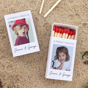 May include: Two white matchboxes with the text "A PERFECT MATCH" and the names "Emma & George". One box is open, revealing red-tipped matches. The other box has a photo of a child wearing a red hat. The boxes are on a sandy surface.