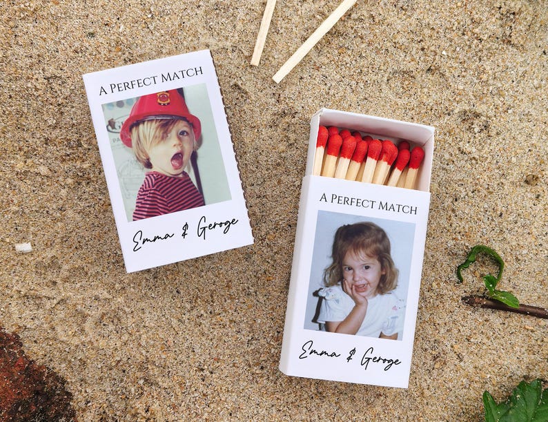 May include: Two white matchboxes with the text "A PERFECT MATCH" and the names "Emma & George". One box is open, revealing red-tipped matches. The other box has a photo of a child wearing a red hat. The boxes are on a sandy surface.