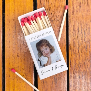 May include: A white matchbox with red-tipped matches, featuring a photo of a young girl and the text "A PERFECT MATCH" and "Emma & George" on a wooden surface. Two loose matches are also visible.