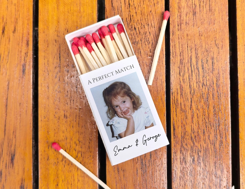 May include: A white matchbox with red-tipped matches, featuring a photo of a young girl and the text "A PERFECT MATCH" and "Emma & George" on a wooden surface. Two loose matches are also visible.