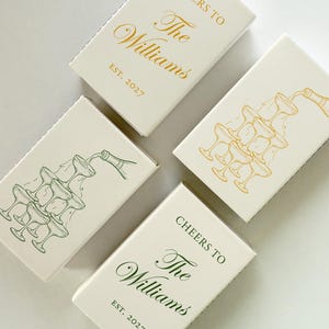 May include: Four rectangular matchboxes with off-white covers. Two feature gold or green line art of a champagne tower. Two feature the text Cheers to The Williams EST. 2027 in gold or green script and serif fonts, matching the champagne tower colors.