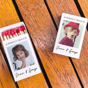 May include: Two white matchboxes with the text "A PERFECT MATCH" and the names "Emma & George". One box is open, revealing red-tipped matches. Each box features a photo of a child. The boxes are on a wooden surface.
