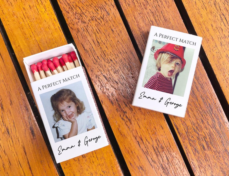 May include: Two white matchboxes with the text "A PERFECT MATCH" and the names "Emma & George". One box is open, revealing red-tipped matches. Each box features a photo of a child. The boxes are on a wooden surface.