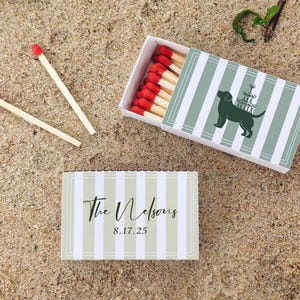 May include: Matchbox with a green and white striped design, featuring a silhouette of a dog with a champagne tower. Two loose matches with red tips are next to the box. The text "The Nelsons 8.17.25" is on a matching box.