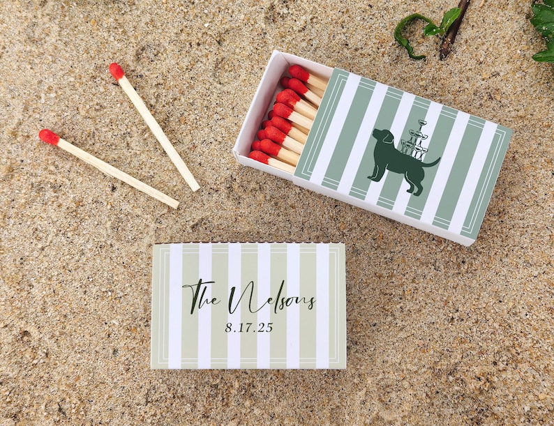 May include: Matchbox with a green and white striped design, featuring a silhouette of a dog with a champagne tower. Two loose matches with red tips are next to the box. The text "The Nelsons 8.17.25" is on a matching box.