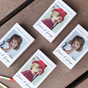 May include: Four matchboxes with a photo of a child, the text "A PERFECT MATCH" and the names "Emma & George" printed on them. The matchboxes are on a brown surface.