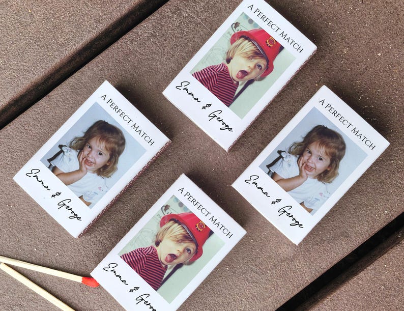 May include: Four matchboxes with a photo of a child, the text "A PERFECT MATCH" and the names "Emma & George" printed on them. The matchboxes are on a brown surface.