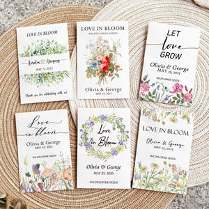 May include: A collection of six white seed packet favors with floral designs and text. The packets feature phrases like "Love in Bloom" and "Let Love Grow," along with names and dates. The packets are arranged on a woven surface.