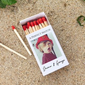 May include: A white matchbox with the text "A PERFECT MATCH" and a photo of a child wearing a red hat. The matchbox also has the names "Emma & George" printed on it. Two matches are lying on the sand next to the matchbox.