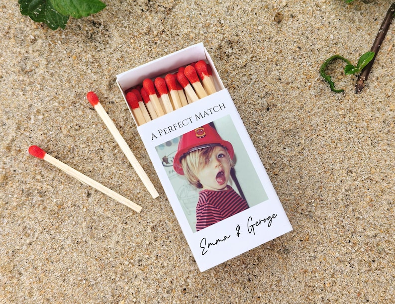 May include: A white matchbox with the text "A PERFECT MATCH" and a photo of a child wearing a red hat. The matchbox also has the names "Emma & George" printed on it. Two matches are lying on the sand next to the matchbox.