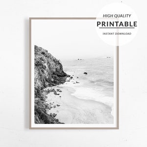 Puede incluir: Fotograf&iacute;a en blanco y negro de un acantilado rocoso que domina una playa de arena con olas rompiendo en la orilla.
