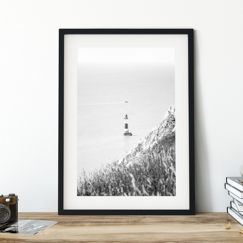 May include: Monochrome framed print featuring a lighthouse with black and white stripes, positioned on a cliff overlooking the sea. A small boat is visible in the distance. The print is displayed on a wooden surface.