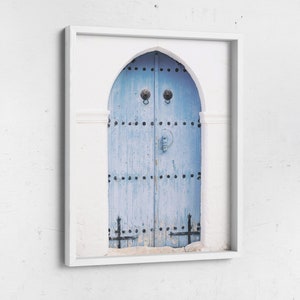 May include: A white framed photograph of a blue wooden door with metal accents and a white archway. The door is weathered and has a rustic look.