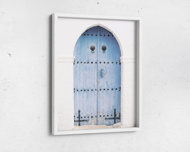 May include: A white framed photograph of a blue wooden door with metal accents and a white archway. The door is weathered and has a rustic look.