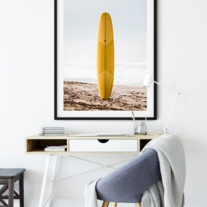 May include: A framed photograph of a yellow surfboard standing upright on a sandy beach. The surfboard is on a beach with waves in the background. The photo is framed in a black frame.