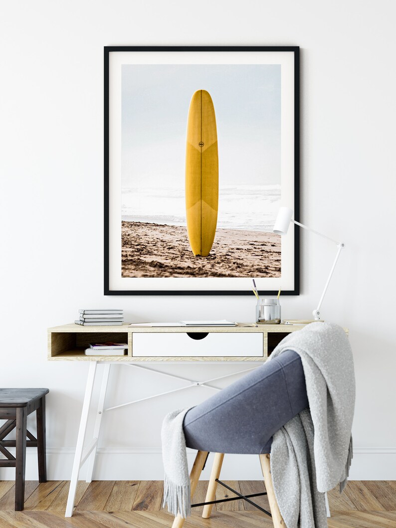 May include: A framed photograph of a yellow surfboard standing upright on a sandy beach. The surfboard is on a beach with waves in the background. The photo is framed in a black frame.