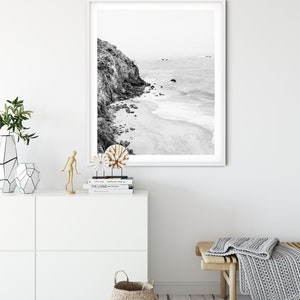Puede incluir: Fotograf&iacute;a en blanco y negro de una costa rocosa con una playa de arena y olas del oc&eacute;ano. La foto est&aacute; enmarcada en un marco blanco y colgada en una pared blanca.