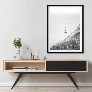 May include: Monochrome framed print featuring a lighthouse on a cliffside. The lighthouse, with black and white stripes, is set against the sea. The print hangs above a wooden console with black legs and a black door.