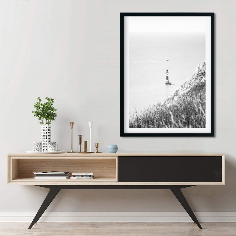 May include: Monochrome framed print featuring a lighthouse on a cliffside. The lighthouse, with black and white stripes, is set against the sea. The print hangs above a wooden console with black legs and a black door.