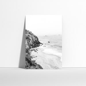 Puede incluir: Una fotograf&iacute;a en blanco y negro de un acantilado rocoso que se encuentra con una playa de arena con olas rompiendo en la orilla.