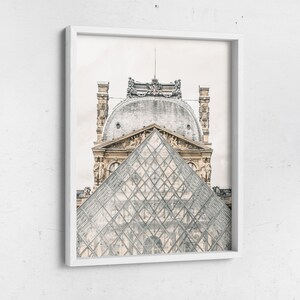 May include: Framed print featuring the Louvre Museum in Paris, France. The image showcases the iconic glass pyramid in the foreground, with the ornate architecture of the museum in the background. The print is encased in a white frame.