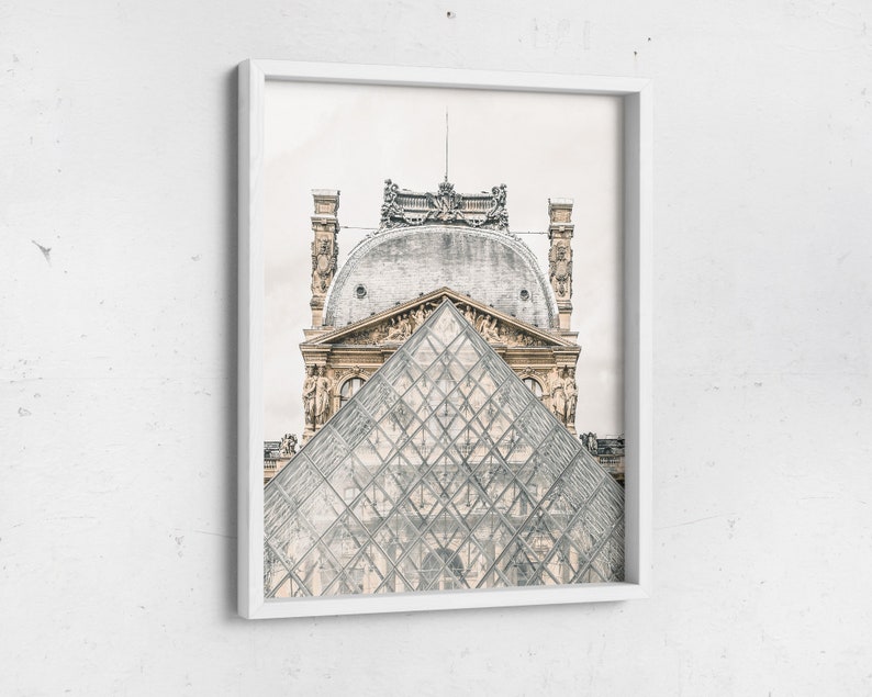 May include: Framed print featuring the Louvre Museum in Paris, France. The image showcases the iconic glass pyramid in the foreground, with the ornate architecture of the museum in the background. The print is encased in a white frame.