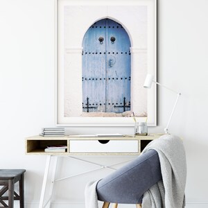 May include: A framed photograph of a blue wooden door with decorative ironwork and a heart-shaped knocker. The door is set in a white wall with an arched opening.