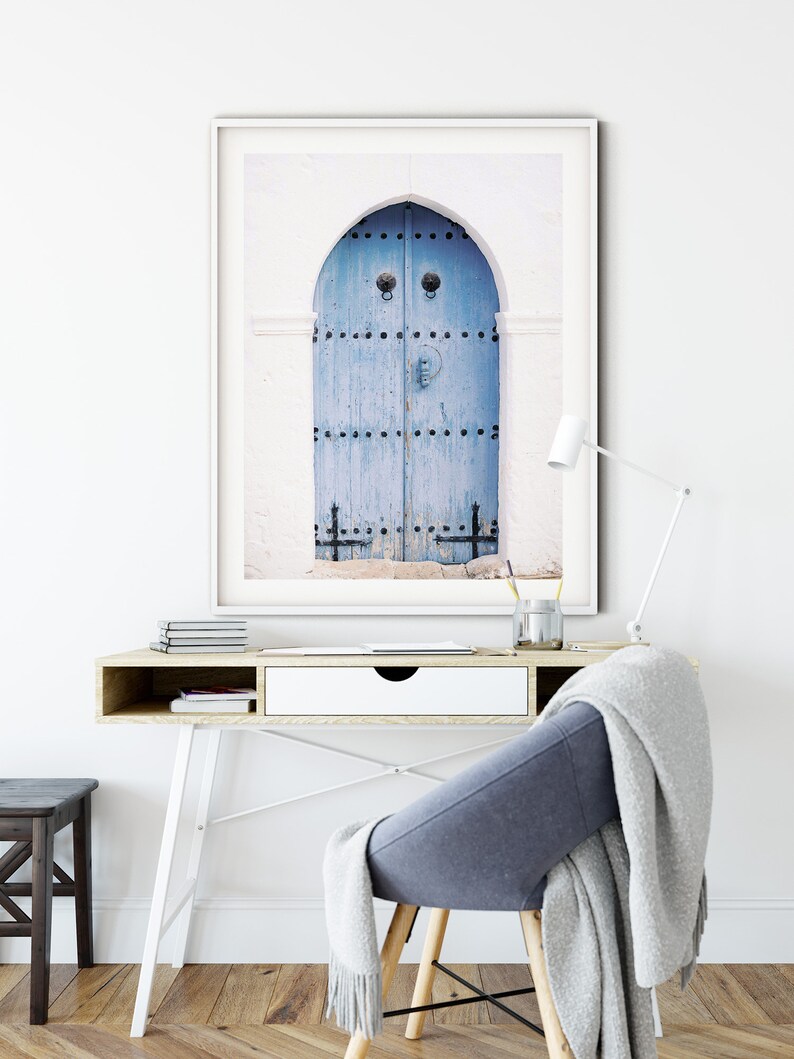May include: A framed photograph of a blue wooden door with decorative ironwork and a heart-shaped knocker. The door is set in a white wall with an arched opening.
