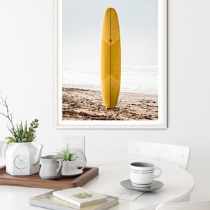 May include: A framed photograph of a yellow surfboard standing upright on a sandy beach. The surfboard has a white stripe down the center and a small circle logo on the side. The beach is empty and the ocean is in the background.