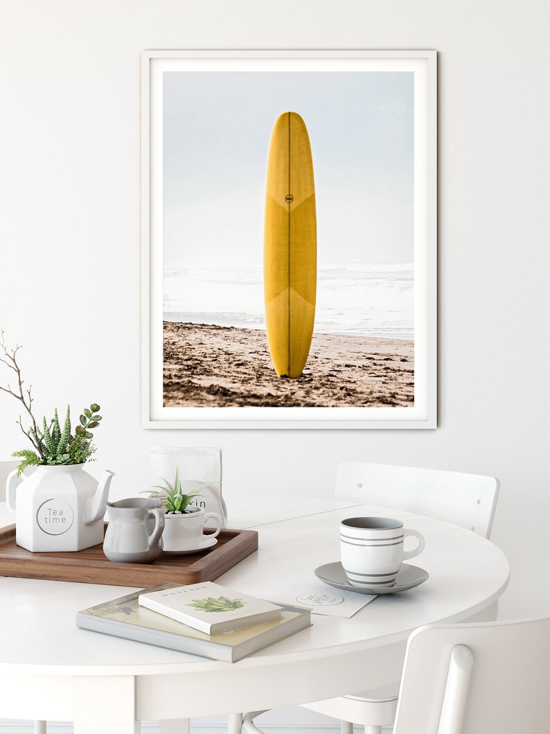 May include: A framed photograph of a yellow surfboard standing upright on a sandy beach. The surfboard has a white stripe down the center and a small circle logo on the side. The beach is empty and the ocean is in the background.