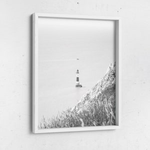 May include: Monochrome framed print featuring a lighthouse with red and white stripes, situated on a small island. A grassy cliff is visible on the right. The sea and sky are a pale grey, creating a serene coastal scene.