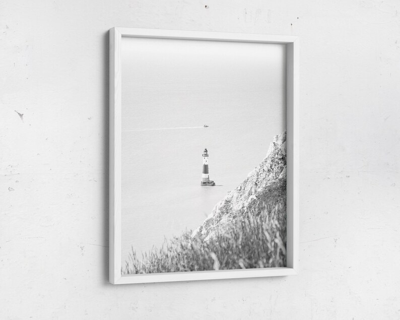 May include: Monochrome framed print featuring a lighthouse with red and white stripes, situated on a small island. A grassy cliff is visible on the right. The sea and sky are a pale grey, creating a serene coastal scene.