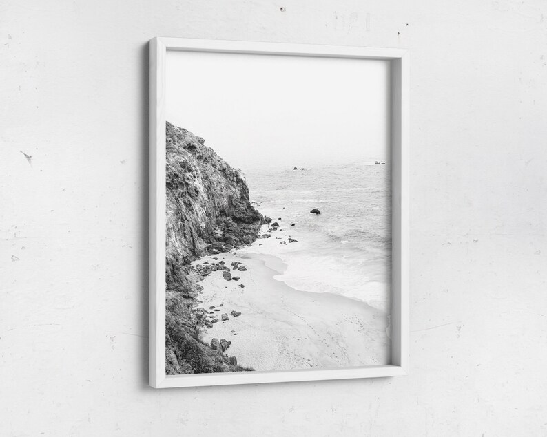 Puede incluir: Fotograf&iacute;a en blanco y negro de un acantilado rocoso que se encuentra con una playa de arena con olas rompiendo en la orilla.
