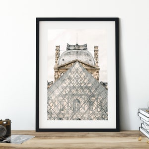 May include: Framed print featuring the Louvre Museum in Paris. The image showcases the iconic glass pyramid in front of the historic building. The print is in a black frame and is displayed on a wooden surface.