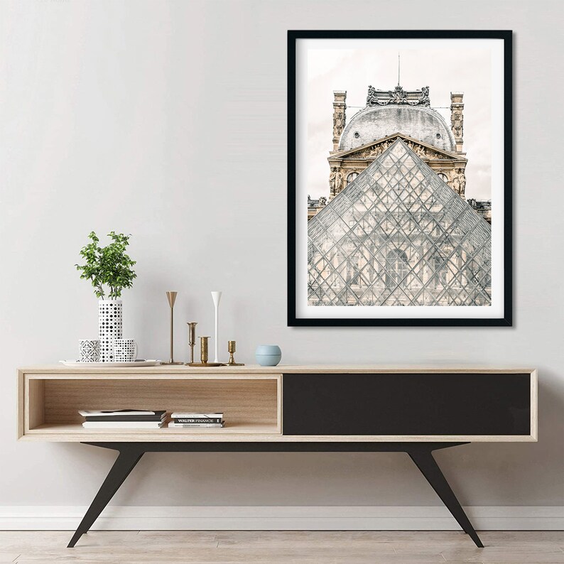 May include: Framed print featuring the Louvre Pyramid in Paris, France. The artwork showcases the glass pyramid in the foreground with the museum's architecture in the background. The print is in a black frame and is displayed above a modern wooden console.