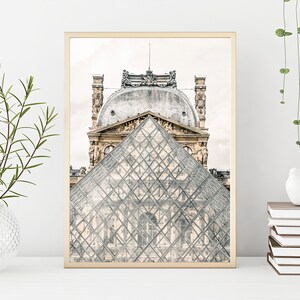 May include: A framed print featuring the Louvre Museum in Paris. The image showcases the glass pyramid in the foreground, with the ornate architecture of the museum in the background. The print has a gold-colored frame and is displayed on a white surface.
