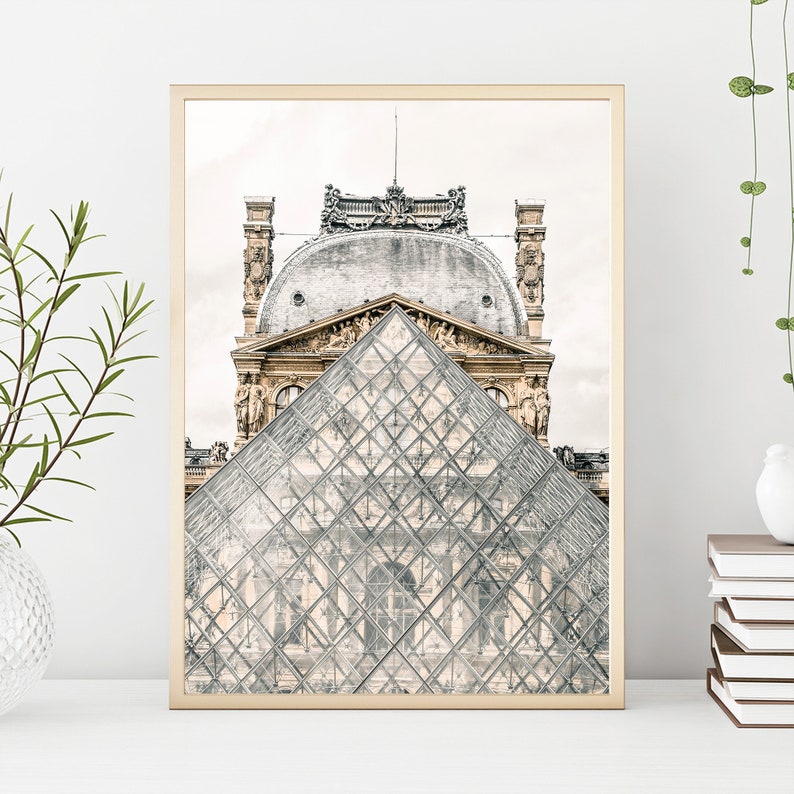 May include: A framed print featuring the Louvre Museum in Paris. The image showcases the glass pyramid in the foreground, with the ornate architecture of the museum in the background. The print has a gold-colored frame and is displayed on a white surface.